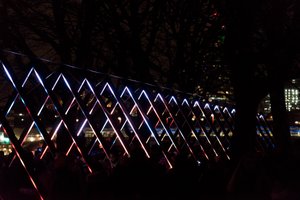 A photo of a light triangular light sculpture. A series of triangles is each lit up in white and bright colours. The triangles recede in to the distance.
