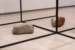 A photo of a sculptural form in a room. There are black struts in a grid shape with two large rocks on the ground.