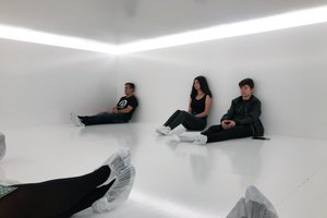 Several people with paper socks are sat in a gloss white room with white light overhead.