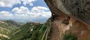 A panorama half looking over the Montserrat countryside and half looking at a path along the side of a mountain that is covered by steep rock.
