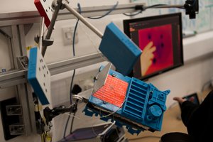 A photo of a science experiment. There are three blue stands held in the air. There’s a screen in the background with a hand visible on it.