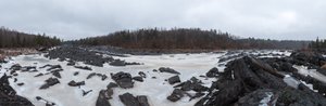 A panorama from the banks of an icy river. There’s dark stones littering the riverbed and creamy ice in between.