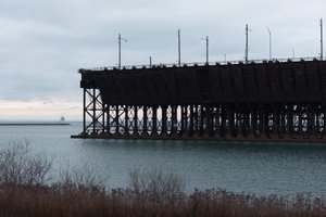 A dark industrial pier in a bay. There’s a small lighthouse in the distance.