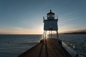 A small lighthouse at the end of a pier is silhouette in front of a setting sun.