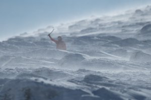 A telephoto photo of Ed on a snowy mountainside. There’s mounds of rocks and snow in the foreground partially obscuring Ed. The camera is focused on the foreground so that Ed is partially blurred.