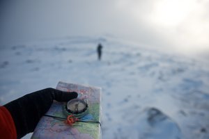 A hand in the foreground holds a compass and map. In the background is a figure walking away. The compass is pointing towards the figure.