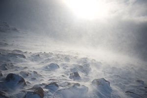 A photo of some snowy rocks on a mountainside. The sun is low in the distance but obscured by snow clouds. There’s flurries of snow all around.