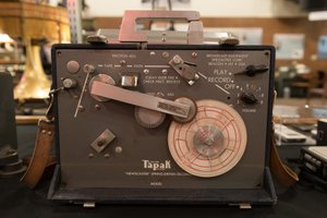 A photo of the front of a vintage recording device. The device has a label on the front reading “Tapak ‘NewsCaster’ spring-driven recorder”