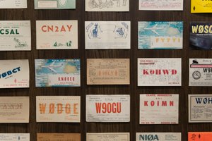 A grid of radio postcards on a wall. The cards are evenly spaced, but each very differently designed. Some have bold typography featuring 5 letter codes. Others depict animals / planes / local scenes.