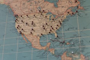 A photo of a map of the United States. There are approximately 30 maroon or grey push-pins inserted at various locations.