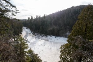 A photo taken from high up, overlooking a bend in Baptism river. The river is mostly covered in snow and ice. There’s sparkles in the air from snow flying about.