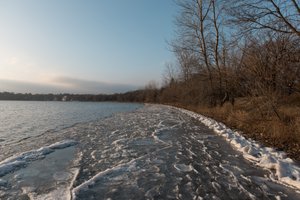 Looking along the shore of Lake Harriet. Close to the shore the edges of the lake have frozen in to intricate shapes. The banks are brown and autumnal.