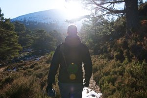 A photo taken from behind as Chris walks ahead. The sun is directly ahead causing Chris to be mostly silhouette with brightly lit edges of his body. The surrounding countryside is fresh with evergreen trees and a snowy mountain in the background.