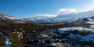 A small stream in a sunny valley landscape. There’s patches of snow on the right bank.