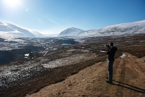 Chris stands on a high path taking a photo of the Gleann Eanaich valley in bright sunlight.