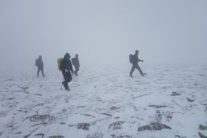 Four people walk on rocky and snowy ground in poor visibility.