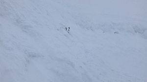A video shot from near the top of Cairngorm mountain. The camera sweeps around a snowy mountain - there’s very little visibility and lots of wind. The camera turns to the right to look inside a snow hole with several people inside.