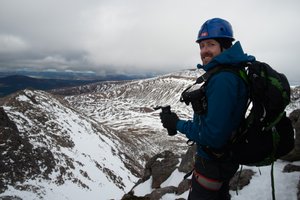 Ed drinking from a vacuum flask, with snowy mountainside in the distance. He’s wearing full climbing gear - helmet, harness, rucksack.
