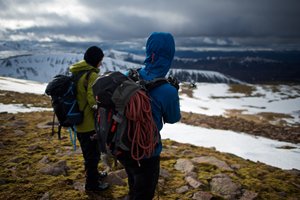 Photo of the backs of Chris and our instructor. Our instructor is pointing downhill with a ski pole. There’s dramatic lighting in the sky - sunlight in the foreground and ominous dark clouds in the background. There are patches of snow on the ground.