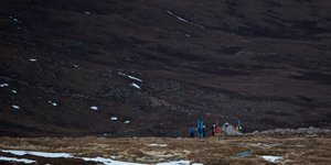 Long shot of several ski mountaineers in a valley. They have skied to the end of the snow, and are now packing up their gear.