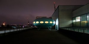 Facing straight on Futuro House. It’s on the roof of a building on a cloudy night.