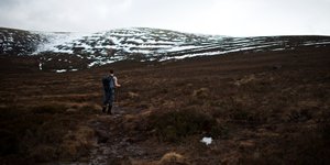 Chris hiking in a t-shirt on a brown grassy path. There’s partially snow covered hills in the distance.