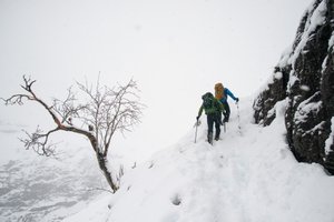 Two hikers climb a somewhat steep snowy hillside. It’s overcast with flurries of snow around.
