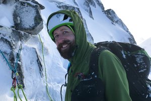 Ed wearing a green softshell jacket with hood over his helmet on the side of a snowy mountain.