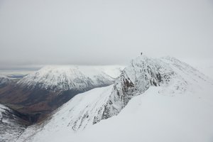 A wide photo of a snow covered mountain peak with other mountains in the far distance beyond a valley. At the top of the peak is a small figure.