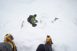 Two boots with crampons on are in the foreground descending a steep snowy mountain. Ed can be seen a few meters ahead.