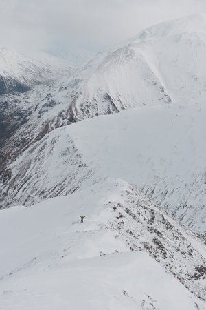 A long photo looking down a snowy mountain to the valley. The small figure of Chris is visible in the distance with both hands in the air waving to the camera.