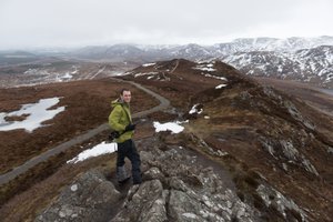 A wide photo looking down on Chris on a mostly brown hillside, with patches of snow here and there.
