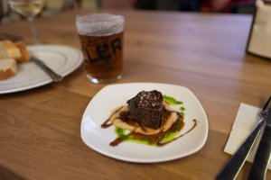 A small piece of beef sits on a plate green vinaigrette and a glass of beer in the background.