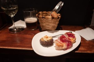A plate of pintxos on a wooden bench next to a basket of bread, a glass of beer, and a glass of white wine. One pinxto is a slice of baguette with ham. The other is half of a sea urchin.