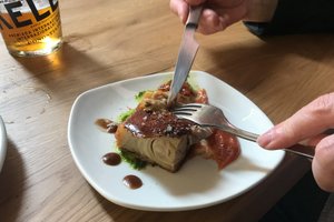 A plate of food just before it gets eaten. A knife and fork are poised to cut in to the plate. The plate contains a single large piece of meat with crispy skin.