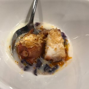 A close up of a small bowl of food with a spoon in the side. The bowl contains two chunks of octopus in a white creamy sauce.