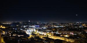 A panorama of Lisbon at night. The streets are filled with yellow and white lights and a suspension bridge can be seen in the distance.