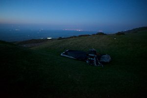 A bivvy bag and rucksack sit in a hollow depression in the grass in the last of the evening light. The sky is dark blue and calm.
