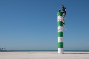 A photo of a small warning beacon on a pier. The beacon is painted in white and green stripes. Ed Horsford is stood on a ladder near the top, looking at the camera.