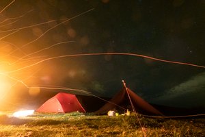 A small tent and a tarp are set up next to each other at night. The camera is close to the ground looking at them, with stars visible in the sky. The left of the frame is bright orange from a nearby fire. There’s trails of sparks crossing the frame from the fire.
