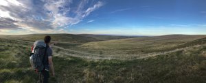 A panorama of rolling hills in Dartmoor in late evening sunlight. On the left a hiker (Oliver Wooderson) with large rucksack points downhill.
