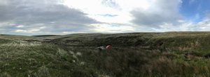 A panorama of a grassy hillside. In the centre a small tent and tarp can be seen set up.