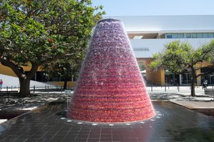 A large fountain in the centre of the frame on a sunny day. The fountain is made from a large circular cone shape, purple-red at the top, fading to red at the bottom.