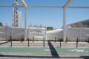 Looking sideways at a road and bicycle path. In the background a white fence blocks the view, but is partially broken revealing some tall grass behind.