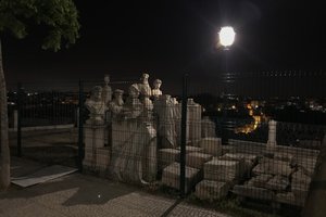 A group of tall marble statues are arranged in a tight grid and surrounded by tall fencing. They’re lit from one side by a streetlamp.