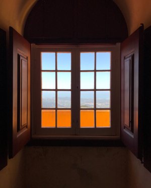 Looking from inside to a window looking out on a high vista. The interior is dark with dark wood window frame. Through the window the sky outside is bright. The window is made from two sets of 2x4 small glass frames. The bottom row is bright orange from an balcony outside.
