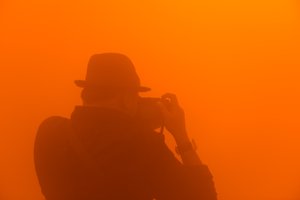 A photographer wearing a hat composes a photo inside an orange foggy room.