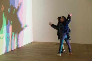 Two people hold hands in a room. A light shines on them from one side, casting a multi-coloured shadow of them against the wall.