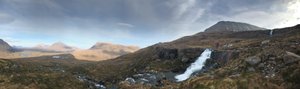 A panorama from the base of Beinn Eighe, looking north. In the right of the frame a small waterfall flows left. Chris is stood at the top of the waterfall in a bright red jacket.