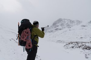 Chris before base of Ben Nevis. He’s got a climbing rope on and axe on the back of his bag, and he’s using a walking pole to point up at the summit.
